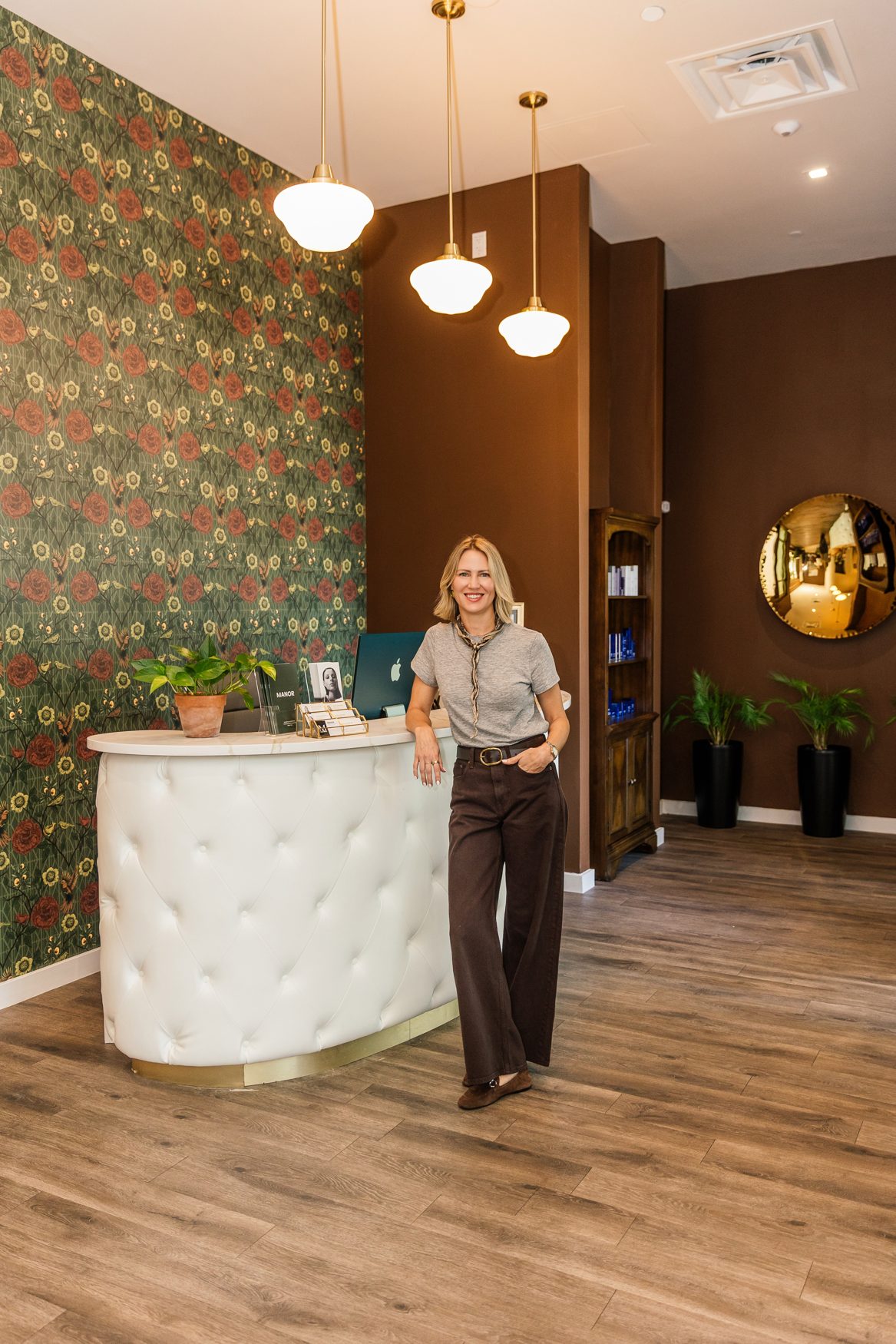 Lindsey Strelecki, BSN, RN at the reception desk of Manor Aesthetics & Wellness in Fort Worth, with the lobby's floral wallpaper, brass pendant lights and product shelves behind her